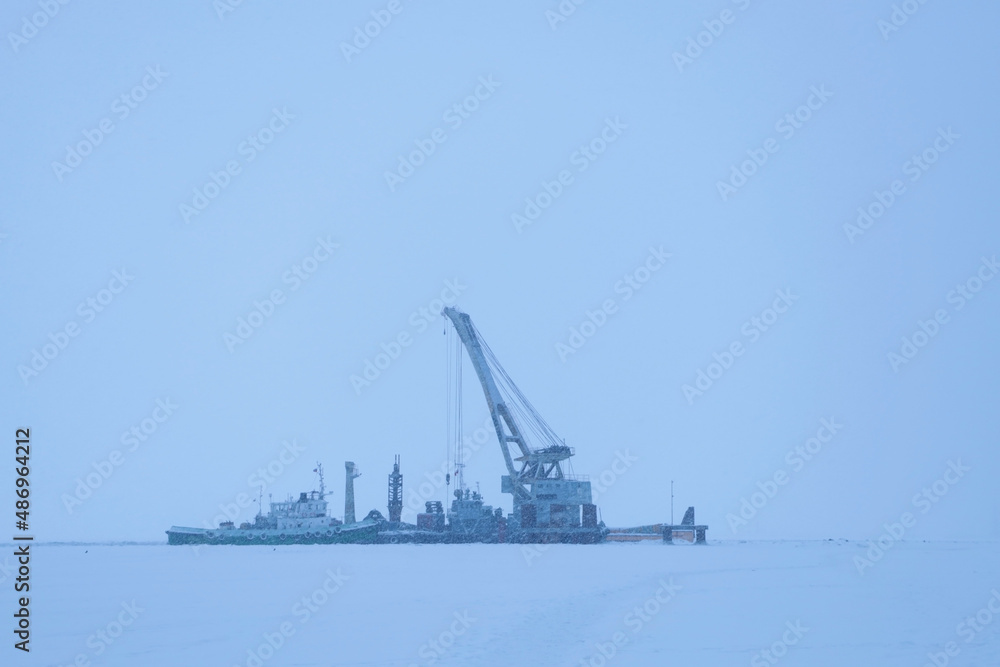 Floating crane for loading and unloading and installation work moored in winter in a frozen bay. Snowfall.