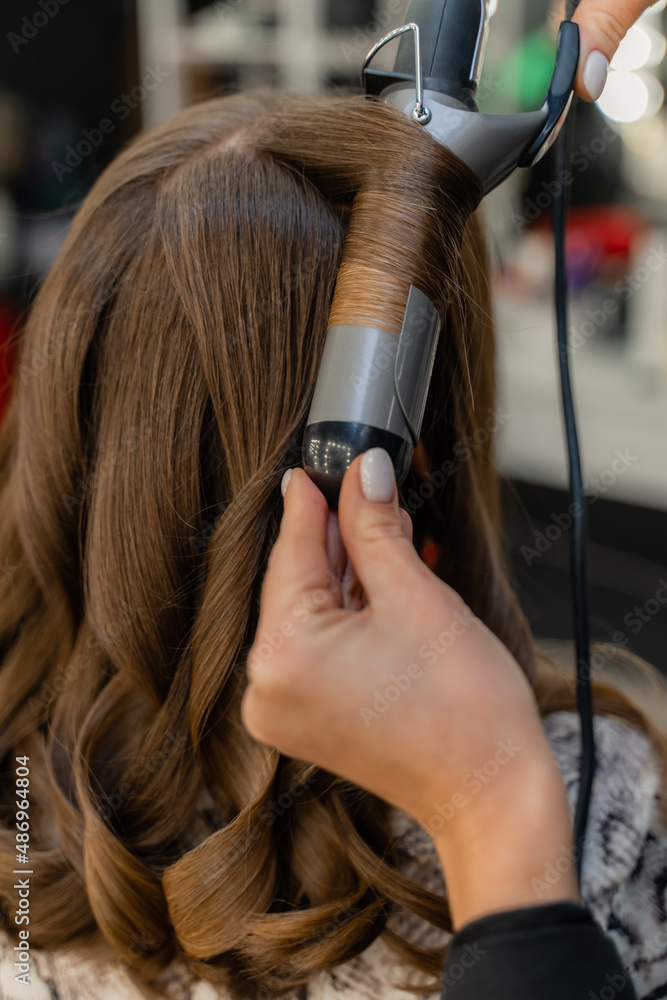 Fototapeta premium The hairdresser prepares a woman, makes a hairstyle with curls using a curling iron. Long light brown natural hair. Self care concept.