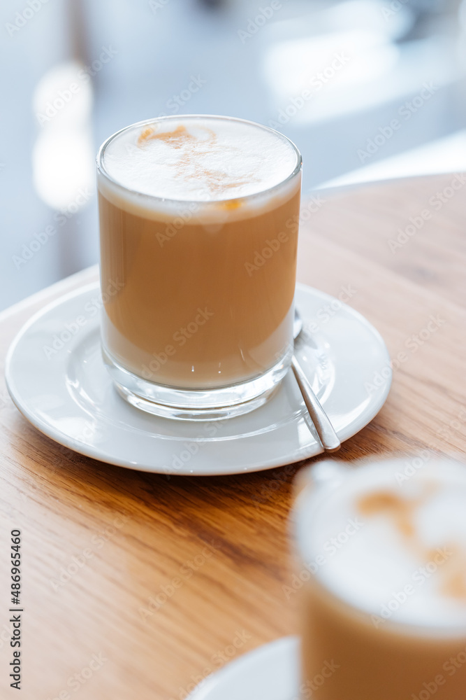 Caffe latte in transparent mug with other mug in first plan, blurred. Barista coffee for international barista day. Break and pause to chill. Coffee with milk and foam. Caffe latte in transparent mug with other mug in first plan, blurred. Barista coffee for international barista day. Break and pause to chill. Coffee with milk and foam.