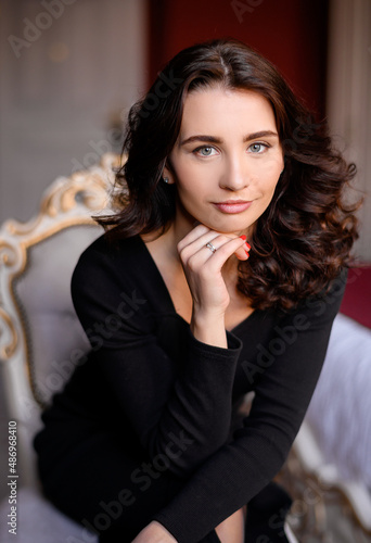 Portrait of sexual woman with beautiful blue eyes and curly hair, which wearing in monochrome black dress, sitting on comfortable chair, keeping hand near face and lovely posing at camera