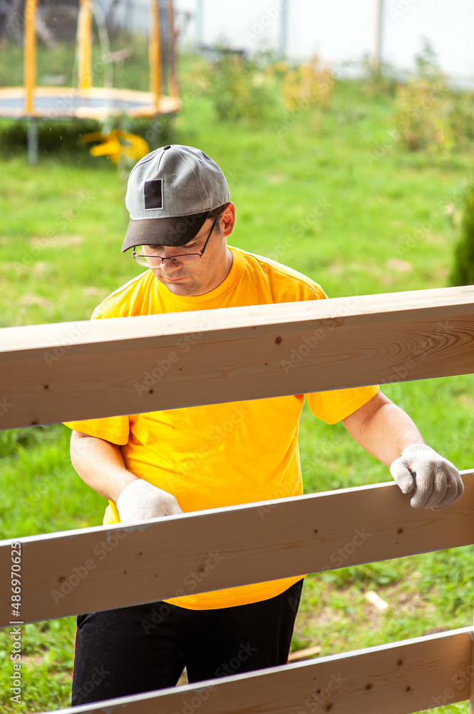 A man in a bright yellow T-shirt and cap holds a brush in his hand and paints the boards on the porch of the house with white paint A man in a bright yellow T-shirt and cap holds a brush in his hand and paints the boards on the porch of the house with white paint