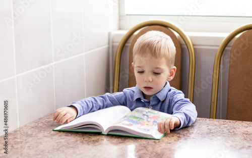 a pensive blond-haired three-year-old boy sitting at the table and reading a book.