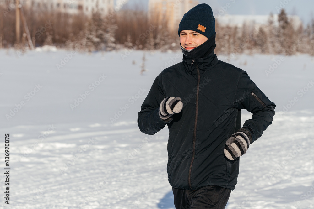 Sporty young man in sportswear jogging through a snowy winter forest. Side view.Playing sports in cold weather. Concept of seasonal sports and acclimatization training