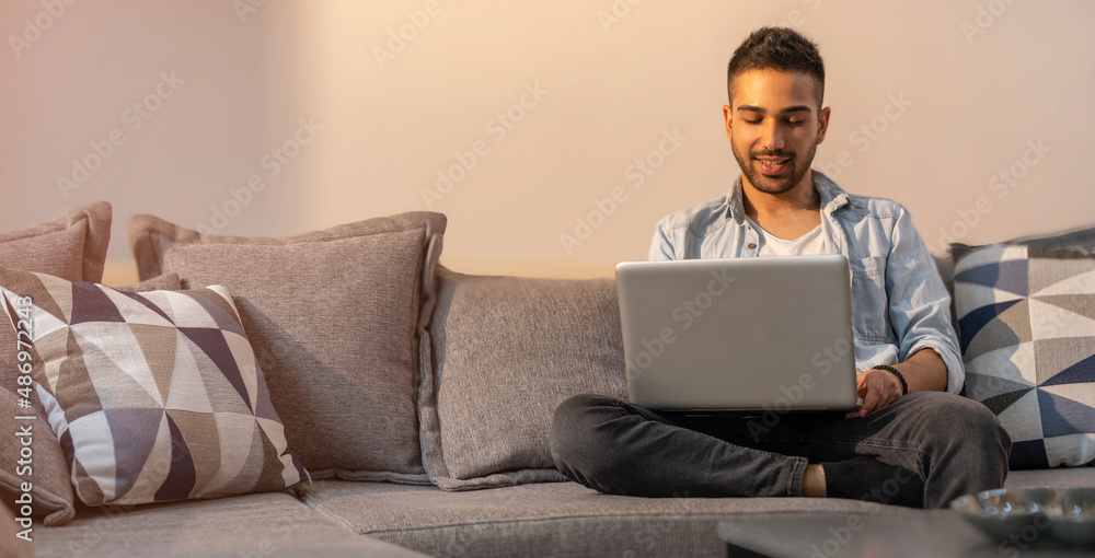Handsome young man using his laptop with smile while sitting on the couch at home. Handsome young man using his laptop with smile while sitting on the couch at home.