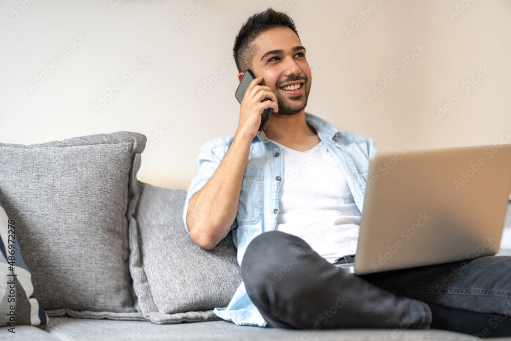Young man talking on mobile phone and using laptop while sitting on couch at home. Young man talking on mobile phone and using laptop while sitting on couch at home.