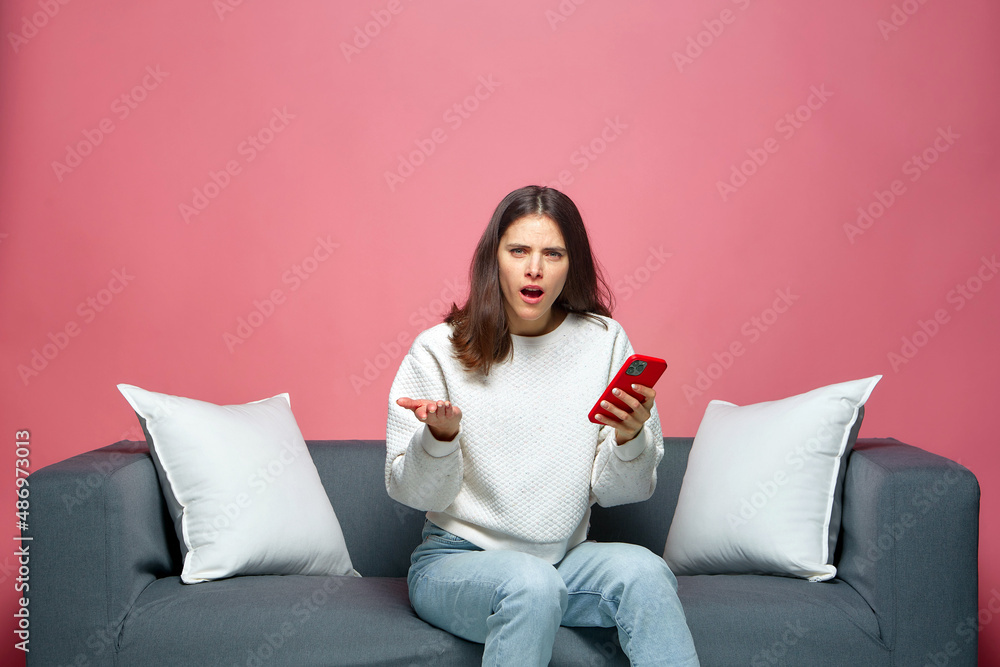Shocked annoyed young woman holds phone irritated by by bad news or spam in message online, sitting on sofa Shocked annoyed young woman holds phone irritated by by bad news or spam in message online, sitting on sofa