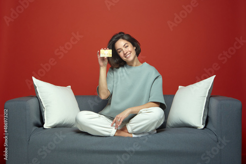 Smiling young woman holding bank credit card with online service, sitting on couch. E-commerce concept