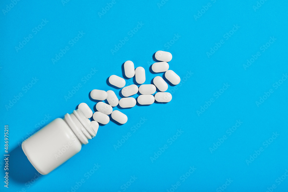 white bottle of pills lies on a blue background. The tablets are scattered side by side white bottle of pills lies on a blue background. The tablets are scattered side by side