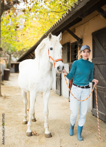 Positive young farmer woman holding reins leading white horse outdoors along stables in farm. Successful horse breeding concept..