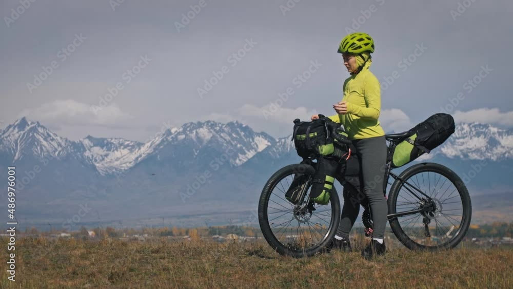 The woman travel on mixed terrain cycle touring with bikepacking. The traveler journey with bicycle bags. Sport tourism bikepacking, bike, sportswear in green black colors. Mountain snow capped. The woman travel on mixed terrain cycle touring with bikepacking. The traveler journey with bicycle bags. Sport tourism bikepacking, bike, sportswear in green black colors. Mountain snow capped.