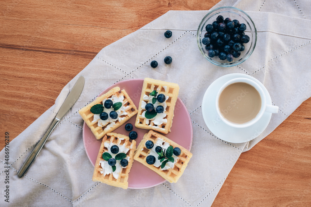 Waffles with cream and blueberries on a plate, a cup of coffee on the table on a cloth napkin. Top view, flat lay Waffles with cream and blueberries on a plate, a cup of coffee on the table on a cloth napkin. Top view, flat lay