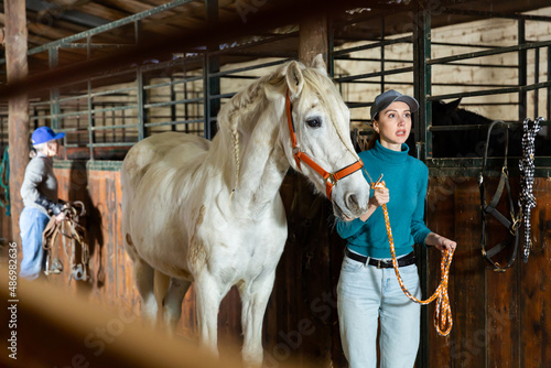 European woman rancher holding rein and leading horse out of stable.