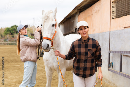 Confident Asian female stable owner holding white racehorse by bridle in outdoor arena while young girl preparing for horseback ride, putting saddle on horse © JackF