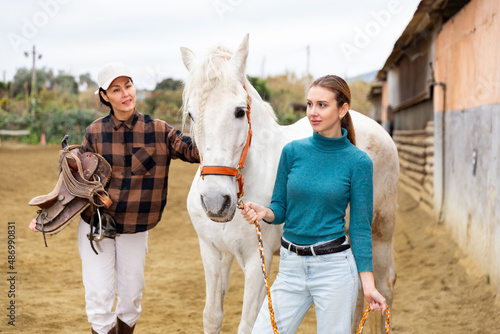 Two women jokey preparing a horse for riding in paddock photo