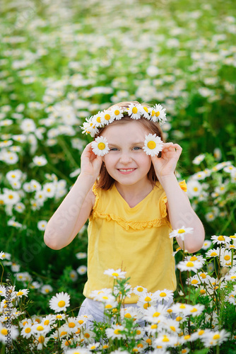 Portrait of a cute village girl in a wreath of daisies in a meadow. He holds a flower in his hands in front of his face. A child on a blooming chamomile field.