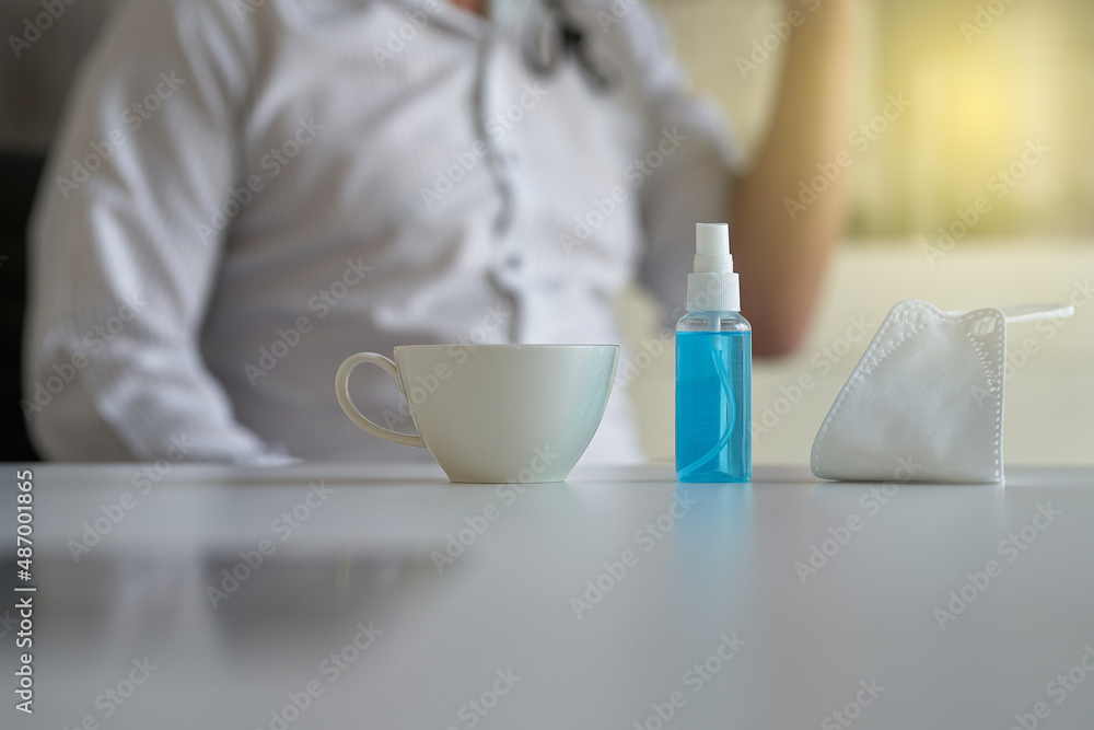 Coffee cup, face mask and hand sanitizer bottle on table in office. Blurred a man worker in office. Coffee cup, face mask and hand sanitizer bottle on table in office. Blurred a man worker in office.