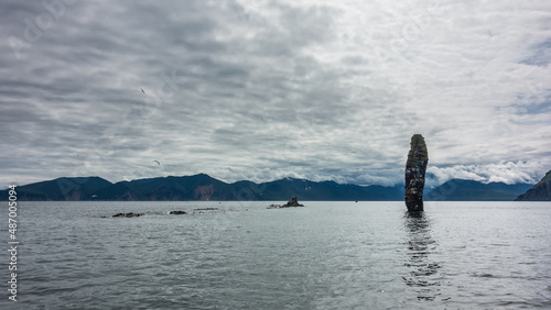 A bizarre picturesque cliff rises above the Pacific Ocean - a high rock that looks like a huge finger. A mountain range against a cloudy sky. Kamchatka. Avacha Bay