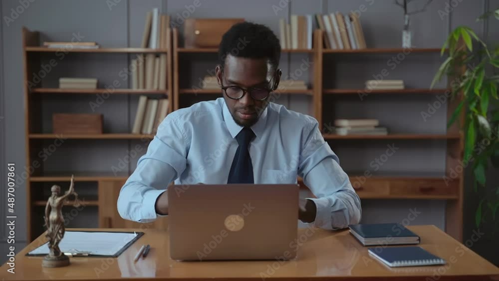 Young man opens laptop and poses while sitting at table in modern office spbas. Portrait of african american businessman takes computer and looks with smile, starts work and sits at desk with statue Young man opens laptop and poses while sitting at table in modern office spbas. Portrait of african american businessman takes computer and looks with smile, starts work and sits at desk with statue