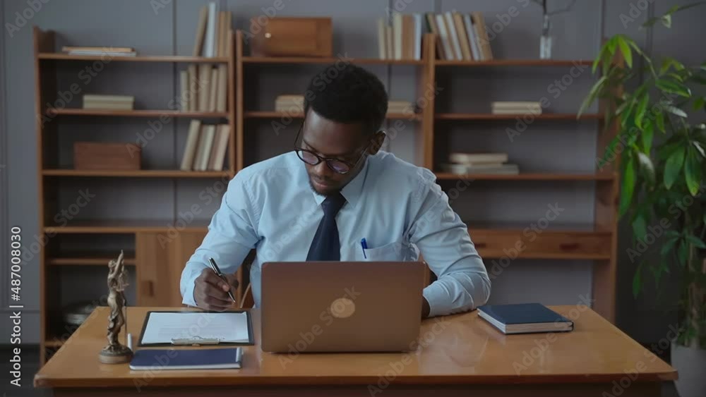 Young businessman using laptop and writing while sitting at table in modern office spbas. Front view of American African man looks at computer screen and writes out terms of contract, does work and Young businessman using laptop and writing while sitting at table in modern office spbas. Front view of American African man looks at computer screen and writes out terms of contract, does work and