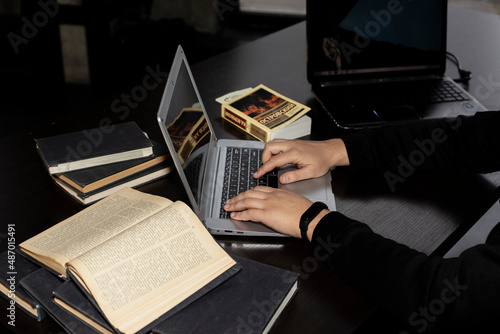There are books and a computer on the table. Male or female hands work on a laptop. A writer or content creator writes text for a magazine, website, or advertisement.