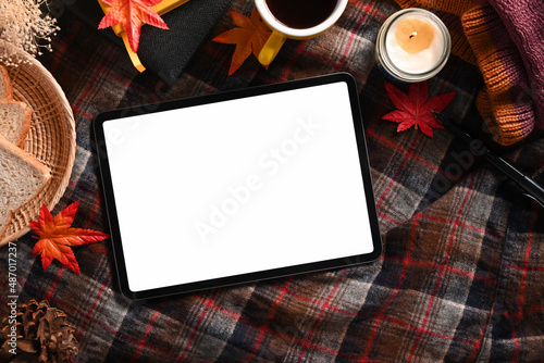 Digital tablet, coffee cup, candle and maple leaves on plaid background.