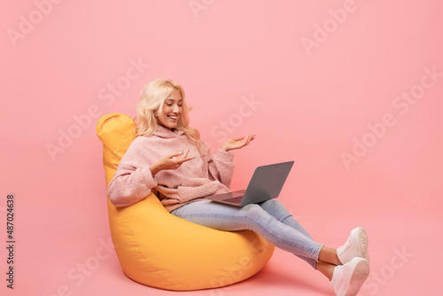 Happy woman making video call on laptop while sitting in beanbag chair, emotionally talking at computer screen