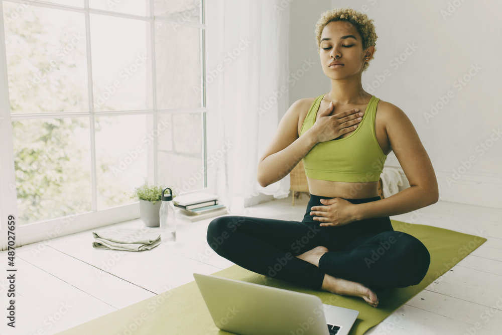 African female yoga teacher having online lesson sitting on green mat in light room showing