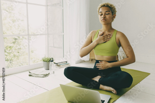 African female yoga teacher having online lesson sitting on green mat in light room showing pranayama techniques, hands on her chest and belly, looking concentrated and focused on body feelings photo