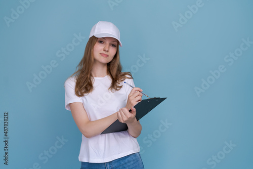 Delivery woman in white uniform isolated on blue background. Girl in white cap and t-shirt, blue jeans, working as a courier or dealer, holding a folder with papers writes. Copy space