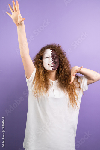 Joyful redhead woman fan with painted face in Qatar flag celebrating on soccer, football match raising one hand up and another hand keeping at shoulder level on violet background.