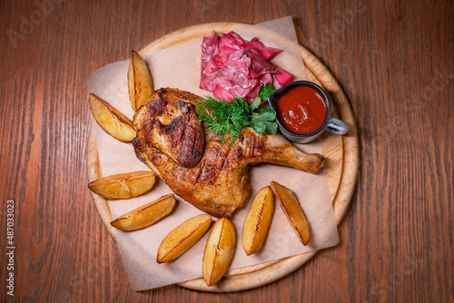 Top view of half of grilled tabaka or tapaka traditional Georgian dish of a pan-fried chicken served with tomato ketchup sauce, roasted potato wedges, fermented cabbage and dill on wooden table photo