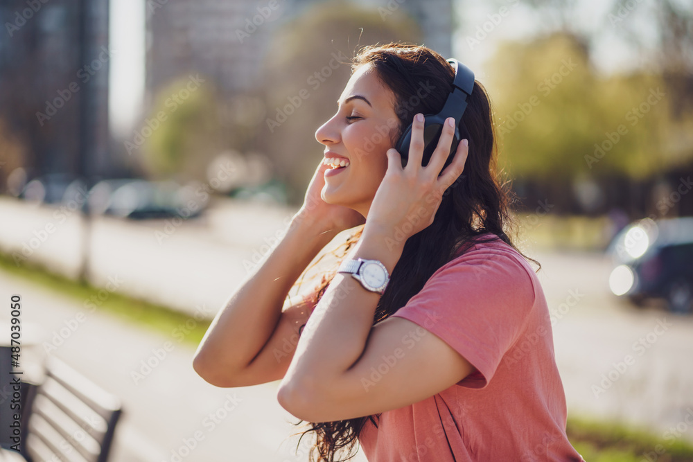Happy young woman is enjoying sunny day outdoor in city. She is listening music on headphones.