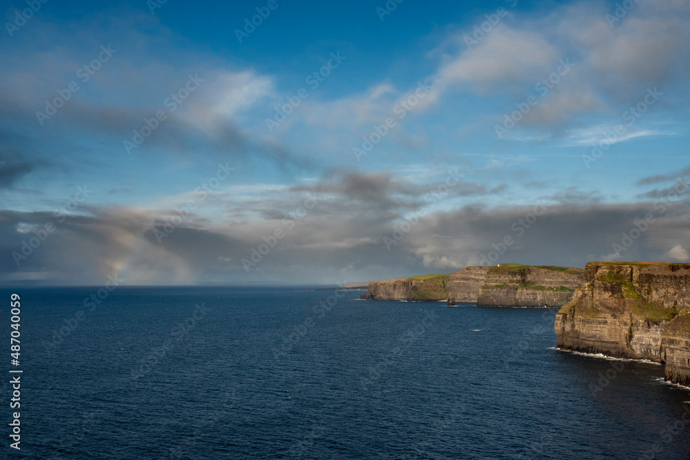 Colorful rainbow in the ocean next to Cliff of Moher, county Clare, Ireland. Amazing scenery with great view. Cloudy sky and blue ocean water. Top Irish nature landmark and travel destination