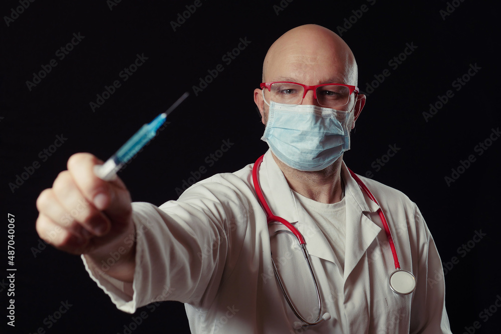 Portrait of a bald doctor with syringe full with blue color vaccine. Dark background. Selective focus. Health care and vaccination concept. Man in white coat, wearing mask and stethoscope. Portrait of a bald doctor with syringe full with blue color vaccine. Dark background. Selective focus. Health care and vaccination concept. Man in white coat, wearing mask and stethoscope.