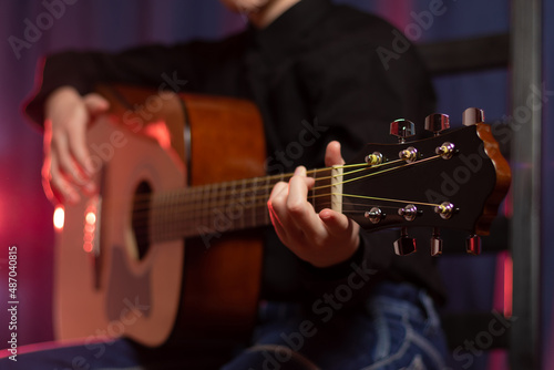 A teenager guy in a black shirt with a guitar in his hands on a purple dark background with red illumination on the stage. Selective focus. Portrait