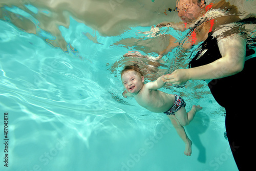 A laughing child with a disability with Down syndrome swims underwater in a children's pool with turquoise water, and his mother holds his hands. Children's disability. Horizontal orientation.