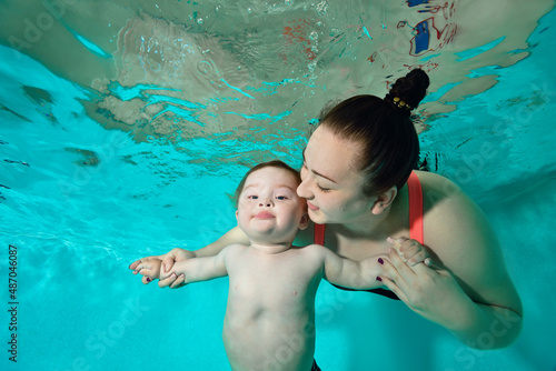 Happy Family: A mother and a child with Down syndrome enjoy swimming underwater in a turquoise water pool. The woman looks at the baby and smiles, holding his hands. Portrait. Horizontal orientation.