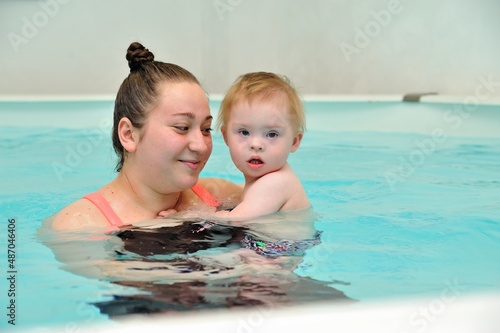 An adult woman holds a baby with Down syndrome in her arms while standing in the water in a children's pool with blue water. She looks at the child and smiles. Portrait. Horizontal orientation.