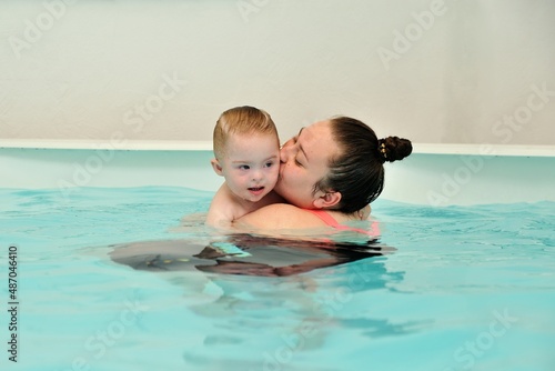 A mother and her child with Down syndrome swim and play in a children's pool with blue water. A woman hugs and kisses her disabled baby tightly. Portrait. Horizontal orientation.