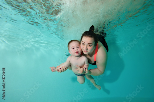 Happy mom and child with Down syndrome dive, swim and pose for the camera underwater in a turquoise water pool. A woman hugs a baby, they look at the camera and smile. Portrait. Horizontal orientation
