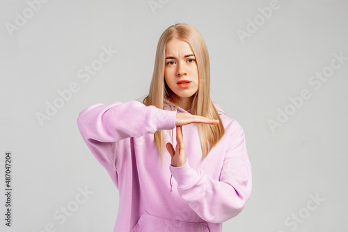 Confused blonde woman standing in trendy hoody over gray background, makes pause or break time gesture, timeout sign, asks to wait, upset about deadline. Body language concept. Time limit photo