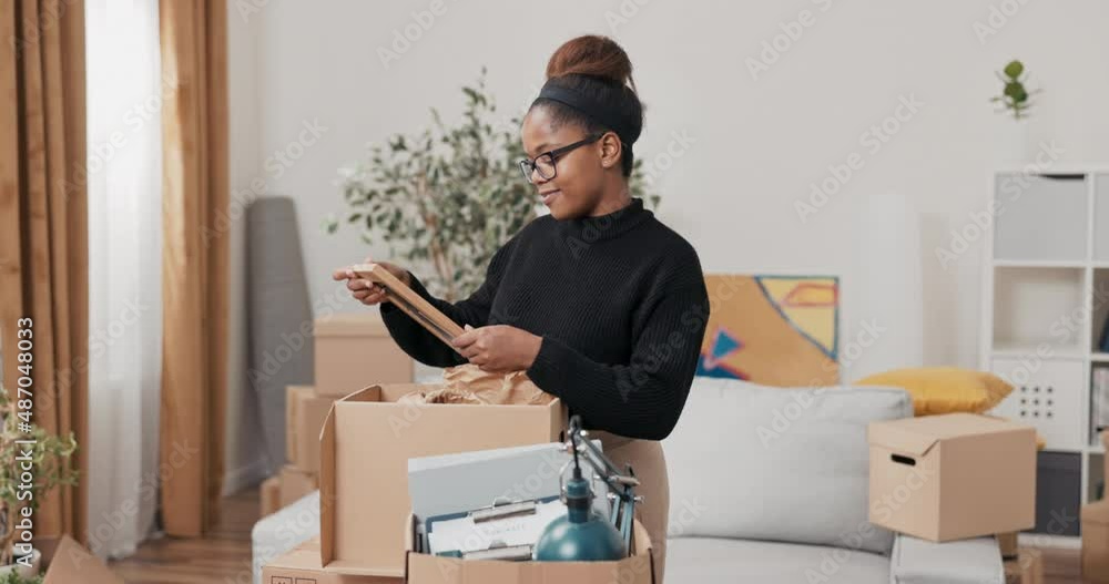 Finding souvenirs during a move, a woman in glasses unpacks a cardboard box in which she finds a picture frame, recalls old times, childhood, loved ones with a smile on her face Finding souvenirs during a move, a woman in glasses unpacks a cardboard box in which she finds a picture frame, recalls old times, childhood, loved ones with a smile on her face