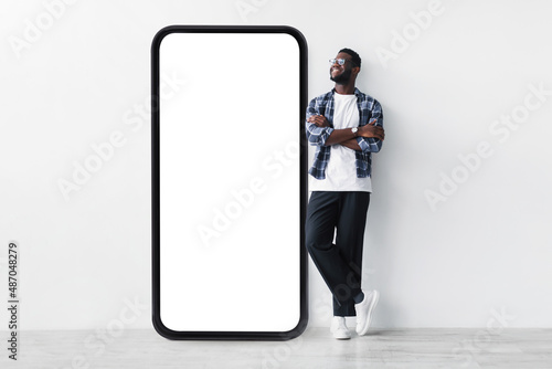 Full body length of black guy standing near huge giant smartphone with empty white screen template, mockup