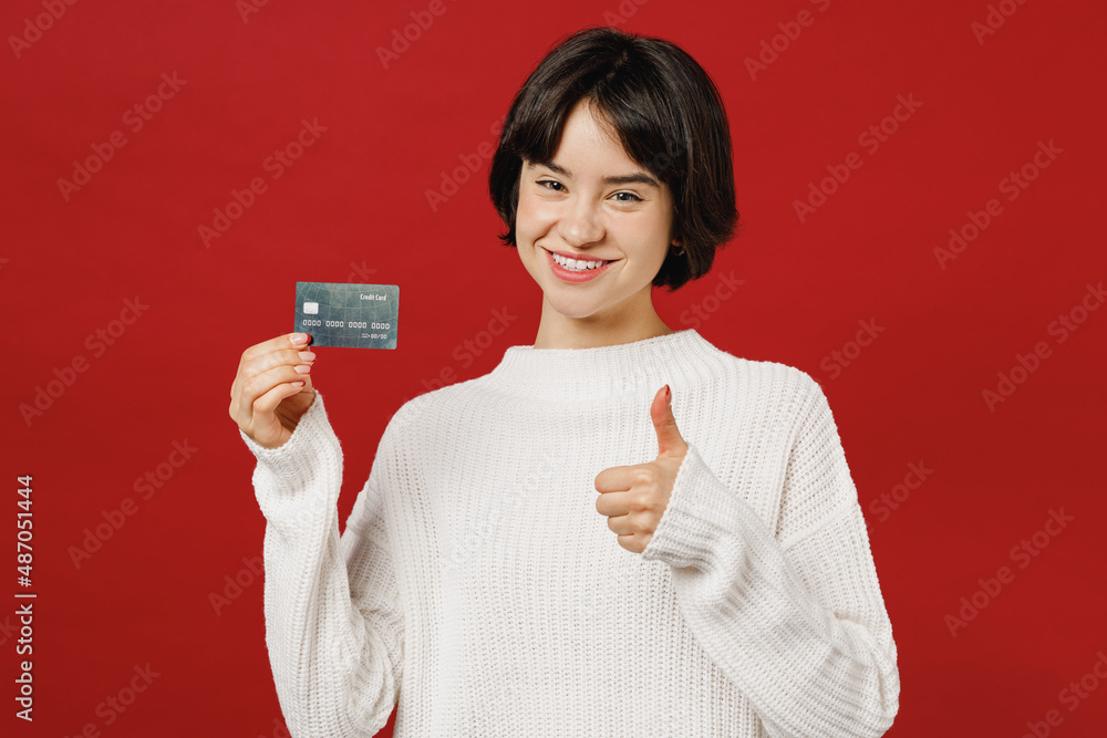 Young cheerful cool smiling happy fun woman 20s wear white knitted sweater hold in hand credit bank card show thumb up isolated on plain red color background studio portrait. People lifestyle concept. Young cheerful cool smiling happy fun woman 20s wear white knitted sweater hold in hand credit bank card show thumb up isolated on plain red color background studio portrait. People lifestyle concept.