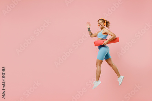 Full body young strong sporty fitness trainer instructor woman wear blue tracksuit headphones spend time in home gym hold caremat isolated on pastel plain light pink background. Workout sport concept.