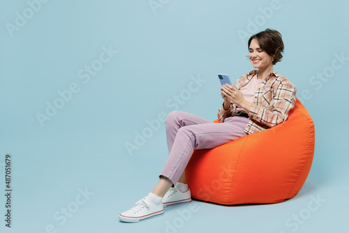 Full body young smiling cheerful happy woman 20s in brown shirt sit in bag chair hold use mobile cell phone isolated on pastel plain light blue background studio portrait. People lifestyle concept