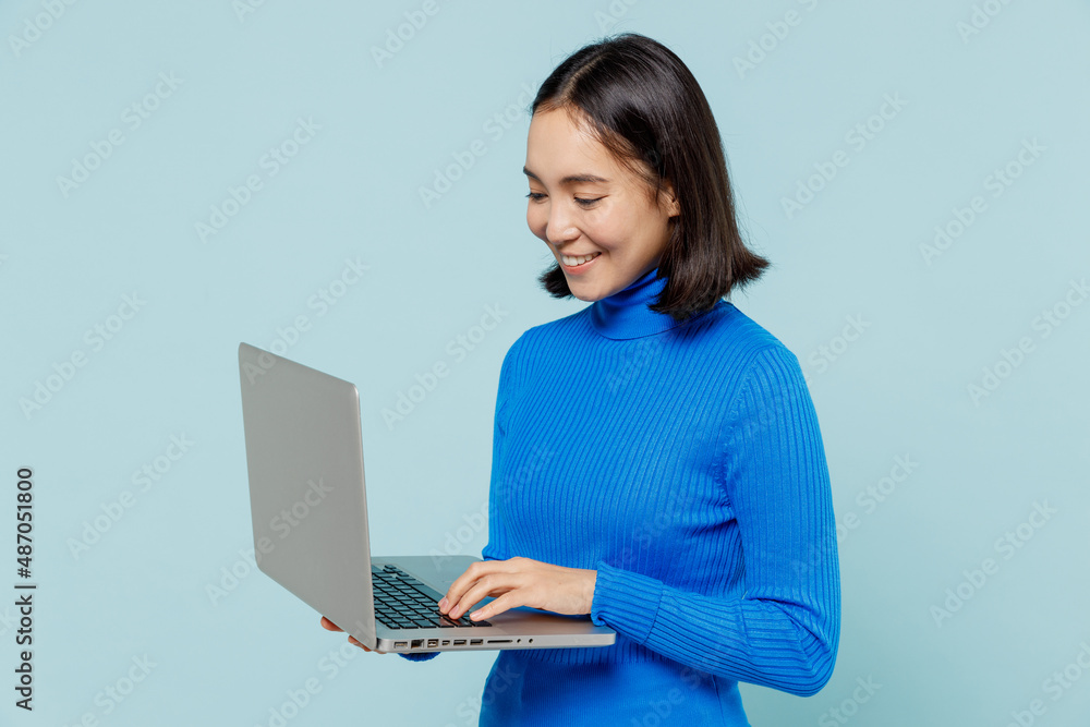 Smiling happy young woman of Asian ethnicity 20s years old wears blue shirt hold use work on laptop pc computer typing browsing chatting isolated on plain pastel light blue background studio portrait. Smiling happy young woman of Asian ethnicity 20s years old wears blue shirt hold use work on laptop pc computer typing browsing chatting isolated on plain pastel light blue background studio portrait.