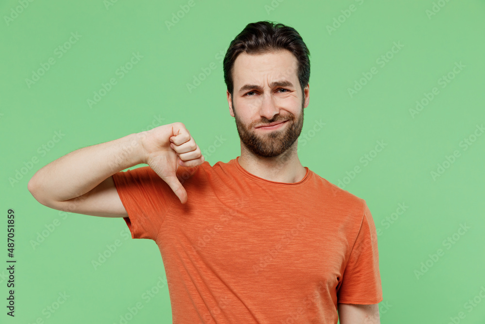 Young displeased disappointed sad man 20s wear casual orange t-shirt showing thumb down dislike gesture isolated on plain pastel light green color background studio portrait. People lifestyle concept. Young displeased disappointed sad man 20s wear casual orange t-shirt showing thumb down dislike gesture isolated on plain pastel light green color background studio portrait. People lifestyle concept.