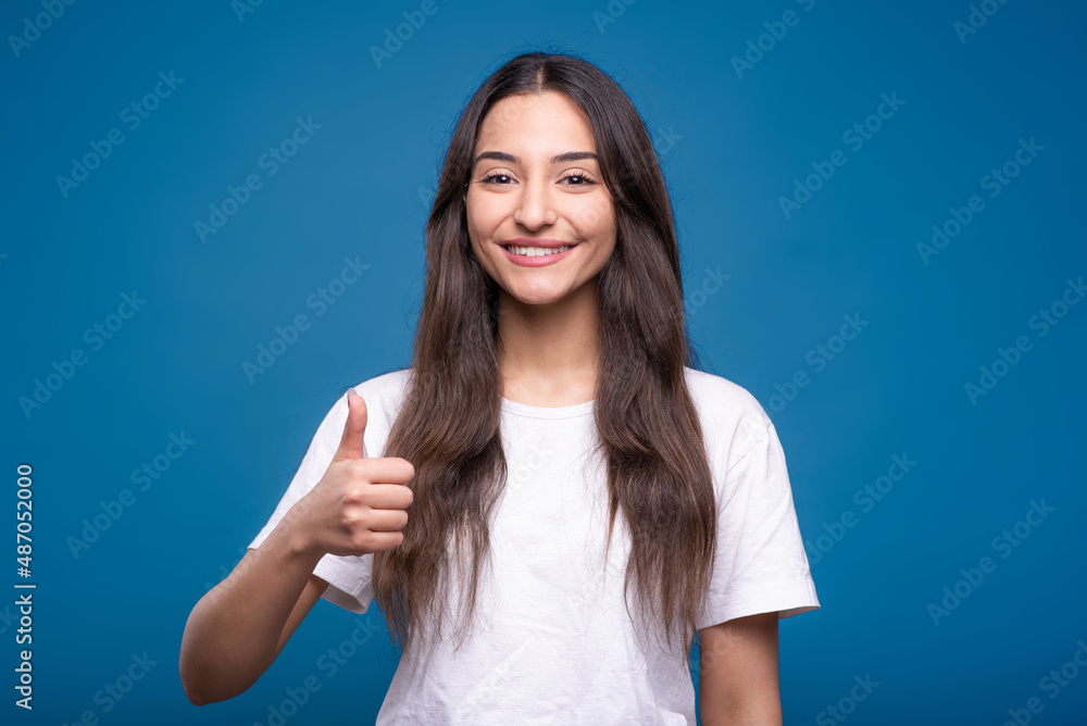 Attractive caucasian or arab brunette girl in white t-shirt showing thumb up and smiling isolated over blue studio background. Attractive caucasian or arab brunette girl in white t-shirt showing thumb up and smiling isolated over blue studio background.