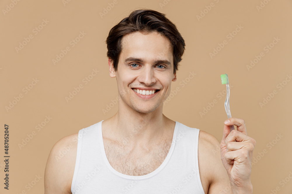 Attractive smiling happy young man 20s perfect skin in undershirt hold brush look camera isolated on pastel pastel beige background studio portrait. Skin care healthcare cosmetic procedures concept. Attractive smiling happy young man 20s perfect skin in undershirt hold brush look camera isolated on pastel pastel beige background studio portrait. Skin care healthcare cosmetic procedures concept.
